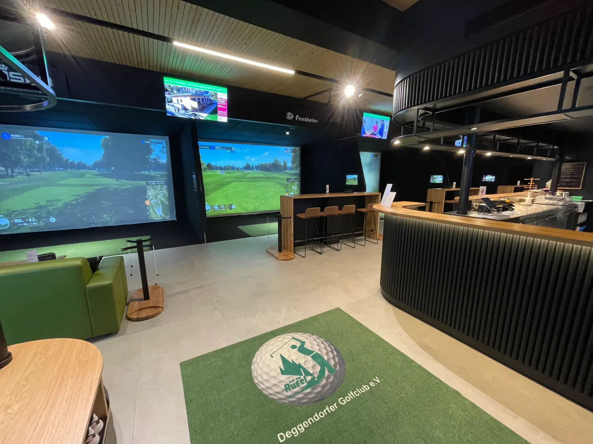 Indoor-Golfhalle der Rusel-Arena: Simulator-Leinwände mit Golfbahnen, daneben die Bar, am Boden ein Teppich mit dem Logo des Deggendorfer Golfclubs